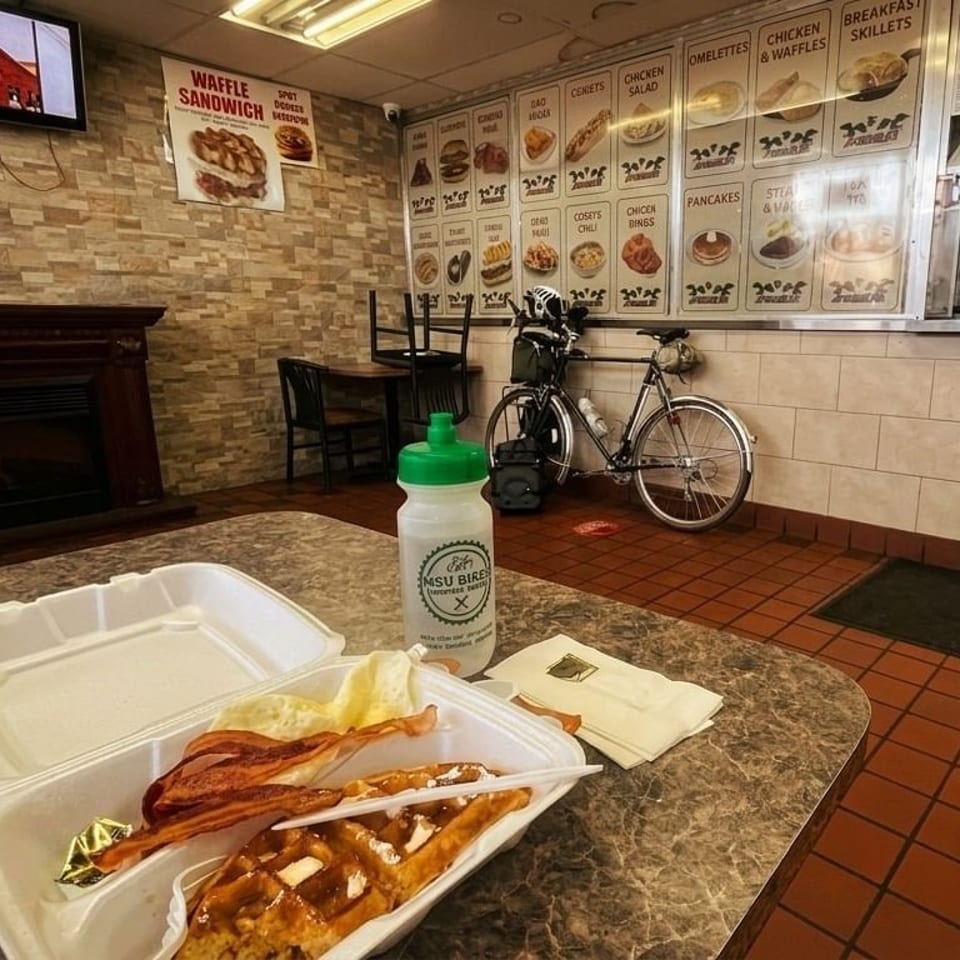 A waffle and bacon in a to-go container on a table inside a diner with menu boards.
